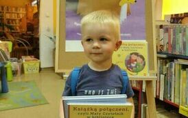 Chlopiec w blond włosach trzyma swoje nowe książki z wyprawki czytelniczej. Za nim stoi tablica z plakatem projektu Mała Książka Wielki Człowiek.