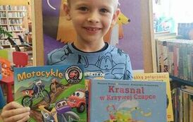Uśmiechnięty chłopiec w blond włosach pokazuje swoją nową książkę i książkę o motocyklach kt&oacute;rą wypożyczył. W tle widać tablicę z plakatem projektu czytelniczego.