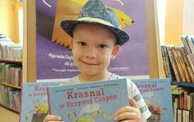 Chłopiec w kapeluszu trzyma w rękach nową książkę z wyprawki, za nim stoi tablica z plakatem projektu.