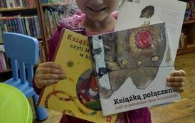 Dziewczynka uśmiechnięta w blond włosach pokazuje swoją wyprawkę czyli dwie ksiązki i kartę z kt&oacute;rej wycina się kostkę do gry.