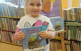 Dziewczynka w ciemnych włosach w bluzeczce Myszka Miki pokazuje swoją nową książkę i zakładkę do książki. Za nią stoi tablica z plakatem projektu.