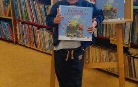 Uśmiechnięty chlopiec w blond włosach pokazuje swoją nową książkę, za nim stoi tablica z plakatem akcji Mała Książka Wielki Człowiek.