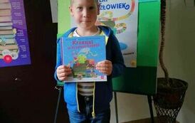 Chłopiec w blond włosach trzyma w ręku nową  książkę, za nim stoi tablica z plakatem akcji Mała Książka Wielki Człowiek.