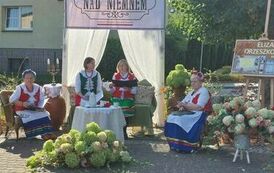 Na placu koło biblioteki siedzą Panie z KGW w strojach kociewskich , jedna z nich czyta tekst "Nad Niemnem".Plac jest pięknie przystrojony w kwiaty i stare meble.