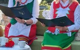 Dwie Panie w strojach kociewskich siedzą przy stoliku z nakryciem kawowym. Jedna z nich czyta tekst " Nad Niemnem". 