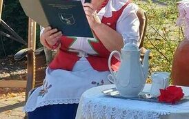 Pani z KGW siedzi przy stoliku z nakryciem kawowym  i czyta tekst o Janie i Cecylii.