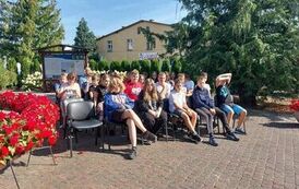 Na placu koło biblioteki siedzi na krzesłach szkolna publiczność słuchając czytania.