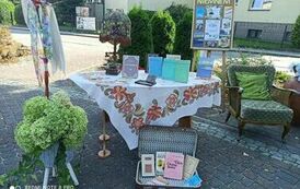 Na placu koło biblioteki stoi st&oacute;ł z lampą i książkami Elizy Orzeszkowej,przed stołem leży walizka z dziełami pisarki. Po prawej stronie stoi tablica korkowa z grafiką "Nad Niemnem" i fotel. A po lewej stronie stoi wieszak na kt&oacute;rym wiszą szaliki.