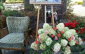 Na placu koło biblioteki stoi tablica korkowa z napisem Eliza Orzeszkowa ze zdjęciami i informacjami. Przed nią stoją kwiaty,a po lewej stronie fotel.