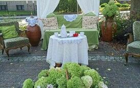 Na placu koło biblioteki wisi baner z napisem "Nad Niemnem". Pod nim są zaczepione białe firanki. Po środku stoi kanapa i  st&oacute;ł z nakryciem kawowym,a przed nim bukiet kwiat&oacute;w. Po obu stronach stołu stoją fotele i wazony w kt&oacute;rych są kwiaty i świecznik.