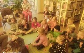 Dzieci siedzą na dywanie i słuchaja bajki, z tyłu widać na krześle opiekuna dzieci.