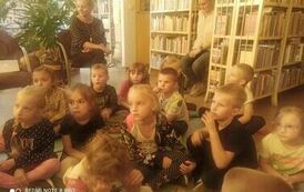 Dzieci siedzą na dywanie i słuchają bajki. Za nimi na krzesłach siedzą opiekunowie.