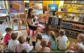 Dzieci siedzą na dywanie, a z przodu widać Panią z biblioteki, kt&oacute;ra cztya im bajkę o jesieni.