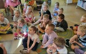 Dzieci siedzą na dywanie i słuchają bajki, kt&oacute;rą czyta Pani z biblioteki.