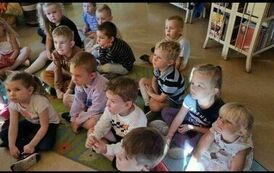 Dzieci siedzą na dywanie i słuchają bajki, kt&oacute;rą czyta Pani z biblioteki.
