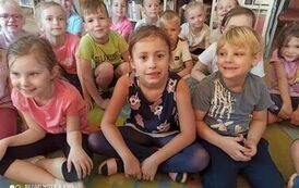 Uśmiechnięte twarze dzieci siedzących na dywanie i słuchających bajki, kt&oacute;rą czyta Pani z biblioteki.