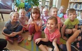 Uśmiechnięte dzieci siedzą na dywanie i słuchają bajki, kt&oacute;rą czyta Pani z biblioteki.