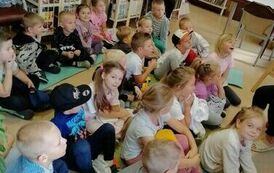Dzieci siedzą na karimatach w bibliotece i słuchają bajki, z tyłu na krzesełku widać Panią opiekunkę.