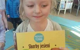 Dziewczynka w blond włosach trzyma książeczkę pt " Skarby jesieni" pokazując ją.