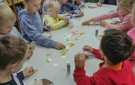 Dzieci stoją przy białym dużym stole i wyklejają z papieu kolorowego jeżyka.