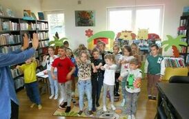 Dzieci stoją na dywanie w rozsypce, z przodu stoi Pani i pokazuje im ruchy taneczne, kt&oacute;re naśladują.