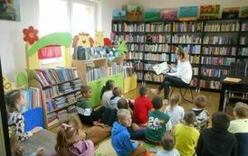 Dzieci siedzą na dywanie i krzesłkach i patrzą w stronę bibliotekarki, kt&oacute;ra czyta im bajkę.