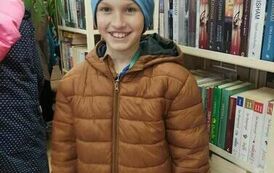 Chłopiec w brązowej kurtce i niebieskiej czapce stoi przy p&oacute;łkach z książkami i się uśmiecha.