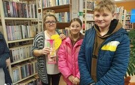 Dziewczynka z chłopcem stoją z bibliotekarką, kt&oacute;ra trzyma w ręku prezent od dzieci.