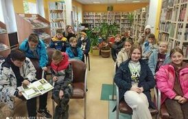 Dzieci siedzą w bibliotece na krzesłach, niekt&oacute;rzy przeglądają książkę. Dzieci siedzą przodem do telewizora.
