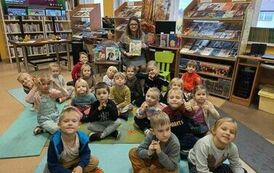 Zdjęcie grupowe dzieci siedzących na dywanie i karimatach, a z tyłu na krzesełku Pani bibliotekarka trzymająca książkę.