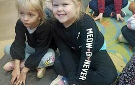 Dzieci siedzą na dywanie i karimatach i słuchają bajki. Z przodu widać uśmiechniętą dziewczynkę w blond włosach. 