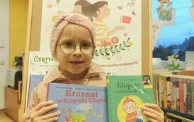 Dziewczynka pokazuje swoją książeczkę z wyprawki czytelniczej i wypożyczoną książeczkę. Za Nią jest tablica z plakatem promującym akcję MAŁA KSIĄŻKA WIELKI CZŁOWIEK.