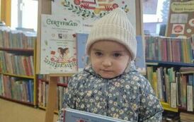 Dziewczynka w jasnej czapce i kurtce w kwiatki pokazuje swoją wyprawkę czytelniczą. Za Nią jest tablica z plakatem promującym akcję MAŁA KSIĄŻKA WIELKI CZŁOWIEK.