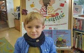 Chłopiec w blond włosach i w niebieskiej kurtce pokazuje swoją książeczkę i zakładkę z wyprawki czytelniczej. Za nim jest tablica z plakatem promującycm akcję MAŁA KSIĄŻKA WIELKI CZŁOWIEK.