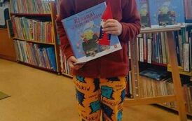 Chłopiec stoi trzymając w rękach książeczkę z wyprawki czytelniczej. Za Nim jest tablica z plakatem promującym akcję MAŁA KSIĄŻKA WIELKI CZŁOWIEK.