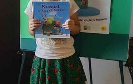 Dziewczynka trzyma w rękach swoją nową książkę z wyprawki czytelniczej.  Za Nią jest tablica z plakatem promującym akcję MAŁA KSIĄŻKA WIELKI CZŁOWIEK.