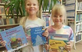 Dwie dziewczynki w blond włosach pokazują swoje wyprawki czytelnicze i wypożyczone książeczki.
