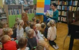Dzieci siedzą na dywanie, a z przodu bibliotekarka opowiada im o książce, w ręku trzyma bajkę o Czarnoksiężniku. Na wprost stoi tablica metalowa obklejona kolorowymi kartkami.