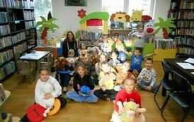 Dzieci siedzą na podłodze, niekt&oacute;re z nich trzymają w rękach misie.Za nimi siedzi ich nauczycielka