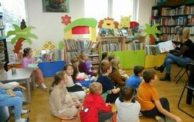 Dzieci siedzą na dywanie i słuchają bajki czytanej przez bibliotekarkę, kt&oacute;ra siedzi z przodu.