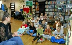 Dzieci siedzą na dywanie i słuchają bajki czytanej przez bibliotekarkę, kt&oacute;ra siedzi z przodu.