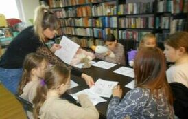 Dzieci siedzą przy stole, przy nich bibliotekarka rozdaje karty pracy.