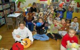 Dzieci siedzą na podłodze, niekt&oacute;re z nich trzymają w rękach misie.Za nimi siedzi bibliotekarka.