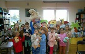 Dzieci stoją w gromadce, niekt&oacute;re  z nich trzyzmają w rękach  małe misie. Z tyłu chłopiec ma uniesione do g&oacute;ry większego białego misia.