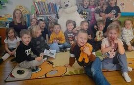 Na środku na krześle siedzi biały duży miś, a wokoło niego siedzą dzieci  na dywanie i ich opiekunowie.