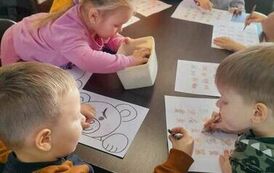 Dzieci siedzą przy stołach i robią karty pracy. Jedna dziewczynka w r&oacute;żowej bluzce szuka w pudełku kredki.