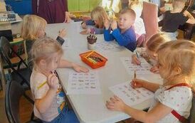 Dzieci siedzą przy stołach i robią karty pracy. Przy białym stole stoi opiekunka i patrzy na ich prace.