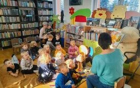 Dzieci siedzą na dywanie i słuchają jak bibliotekarka czyta im bajkę o misiach.