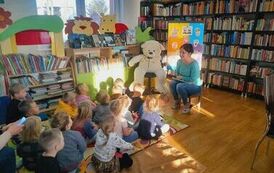 Dzieci siedzą na dywanie i patrzą w stronę bibliotekarki, kt&oacute;ra trzyma w ręku książkę i coś im opowiada. Prz bibliotekarce siedzi biały duży miś i stoi tablica przystrojona misiami z papieru.