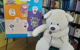 Po lewej stronie stoi metalowa tablica przystrojona misiami z papieru, a po prawej na krześle siedzi duży biały miś.